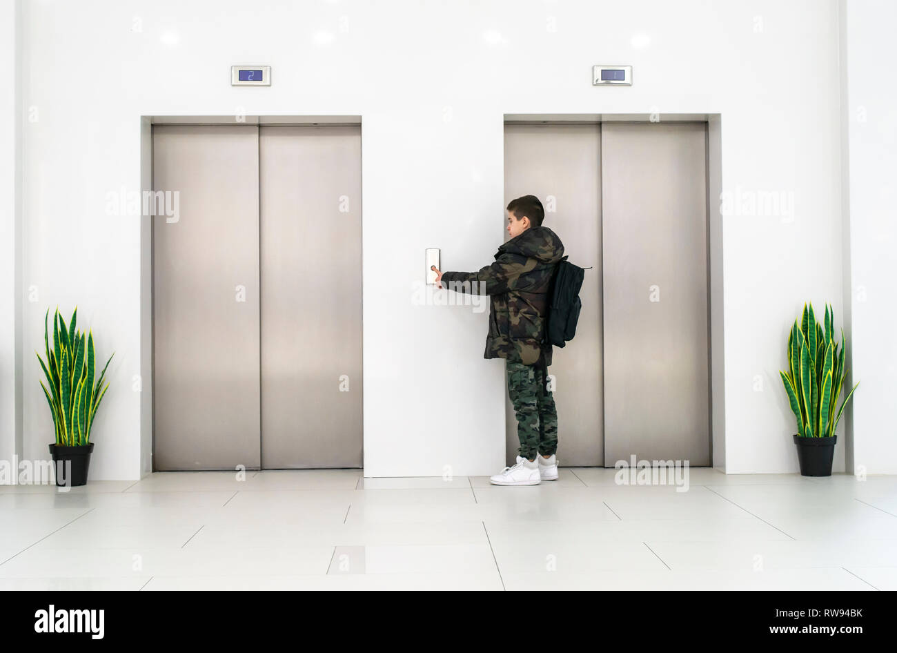 Ragazzo con un abbigliamento informale e white sneakers chiamare l'ascensore. Bianco edificio contemporaneo degli interni. Fiori in vaso e muro bianco. Ascensore metallico Foto Stock