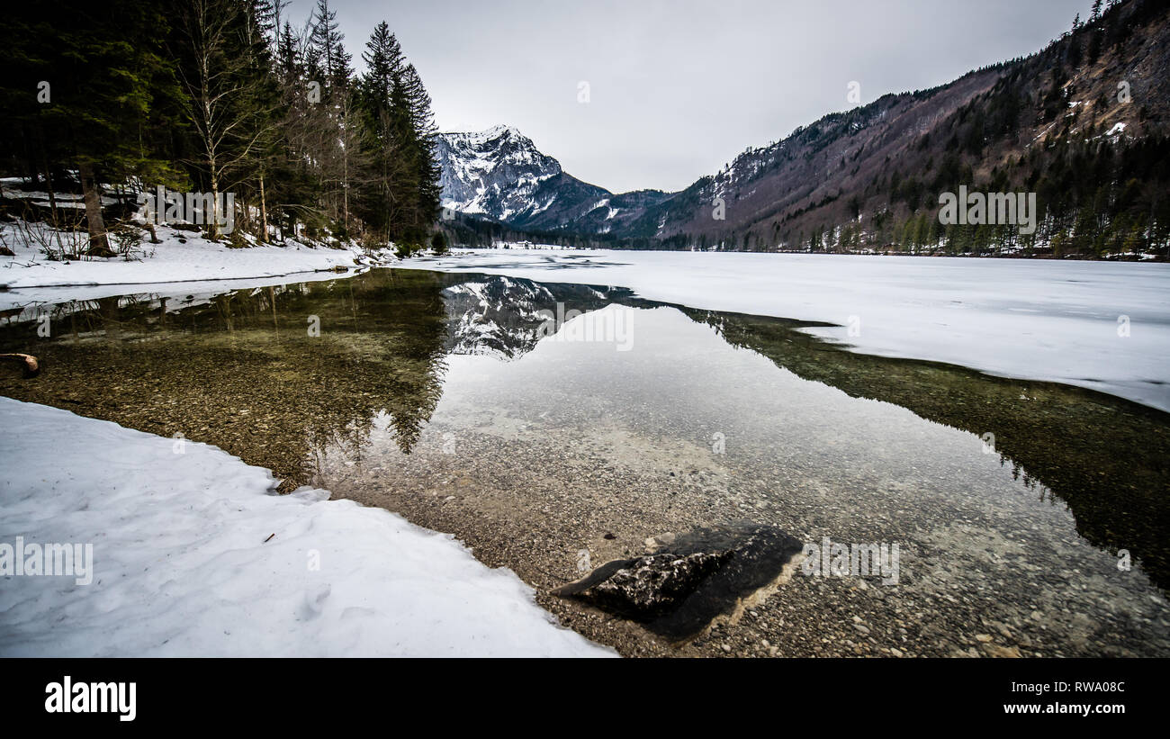 Uno spesso strato di ghiaccio sul lago ghiacciato a Vorderer Langbathsee. Belle montagne dalle vette innevate riflessa nelle acque cristalline del lago Foto Stock