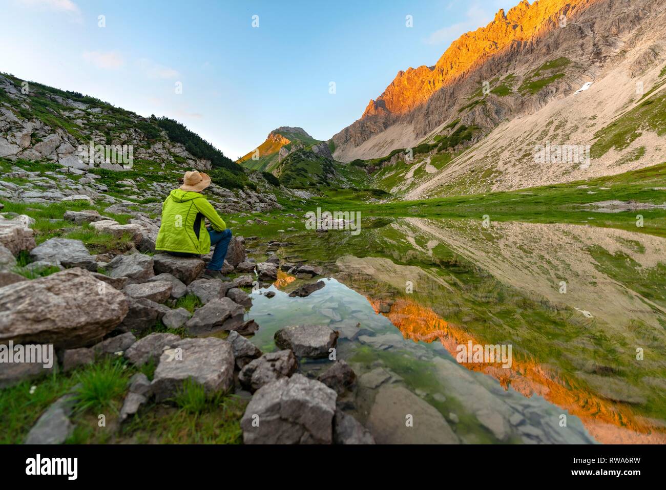 Giovane uomo seduto su di una pietra, la riflessione nel lago di montagna alla Prinz-Luitpold-house, montagne al tramonto, Algovia Alpi Foto Stock