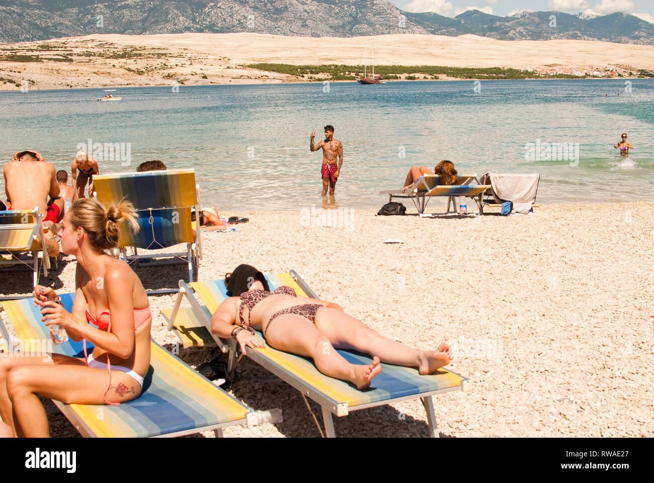 Giovane uomo che indossa pantaloni corti e tenendo selfie in piedi al mare sulla spiaggia di Zrce, con bagni di sole donna in primo piano. La Croazia durante il nascondiglio festival Foto Stock