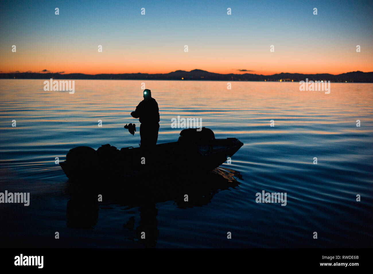 Uomo che indossa un proiettore si erge nel mezzo di una barca e raccoglie in duck decoy mentre galleggiante su un lago come il sole tramonta. Foto Stock