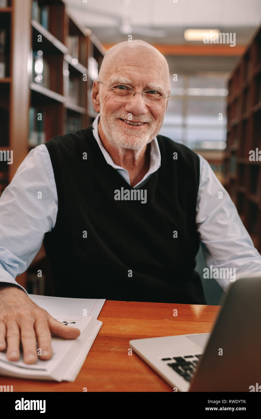 Senior sorridente uomo seduto in biblioteca con libri e computer portatile. Close up di un anziano uomo seduto in una classe e l'apprendimento. Foto Stock