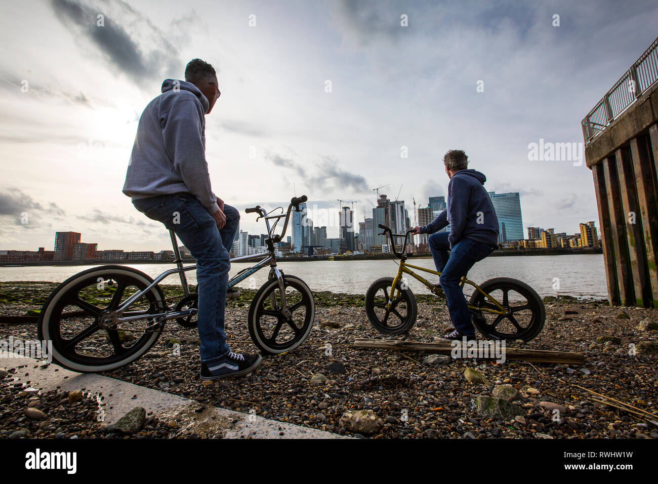 Due uomini su biciclette BMX a Greenwich Peninsula affacciato sul Canary Wharf distretto finanziario attraverso il fiume Thames, London, England, Regno Unito Foto Stock
