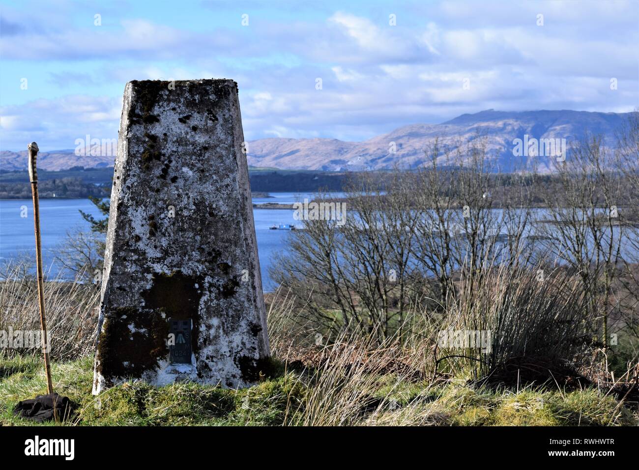 Ordnance Survey pilastro di triangolazione affacciato sul mare scozzese fattorie di gabbie di salmone a Loch Creran, Argyll. Bastone da passeggio mostra proporzioni. Foto Stock