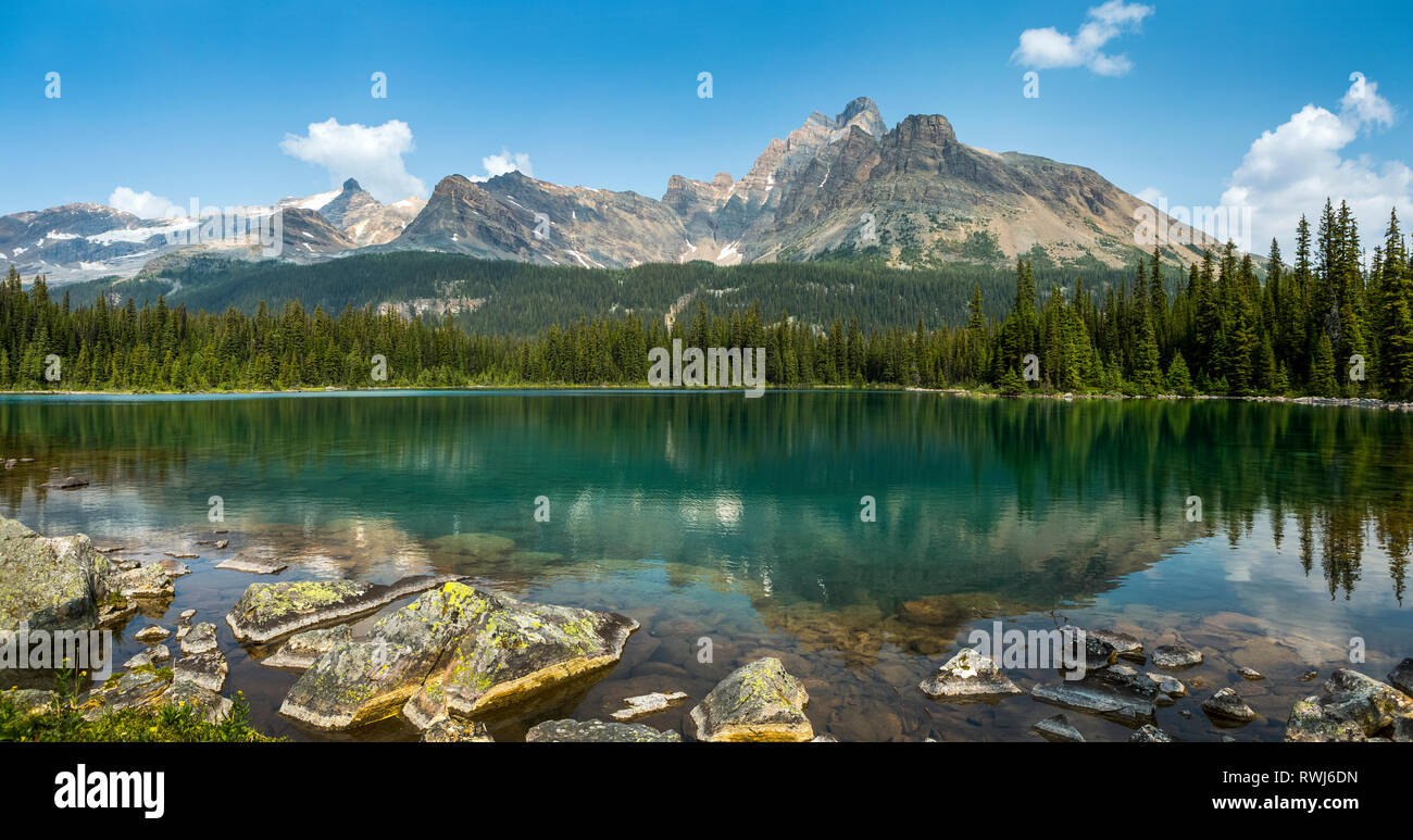 La gamma della montagna riflettendo su un lago alpino con una spiaggia rocciosa e cielo blu e nuvole; British Columbia, Canada Foto Stock