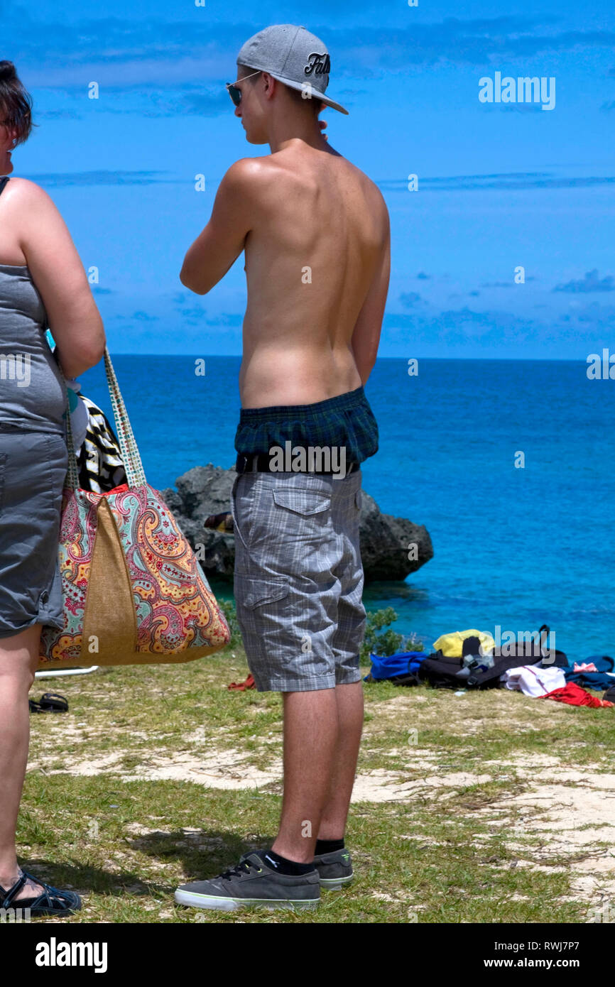 Giovane uomo in piedi; costume da bagno; Pantaloncini metà cadere; acqua; berretto da baseball indietro, Bermuda, estate; verticale Foto Stock