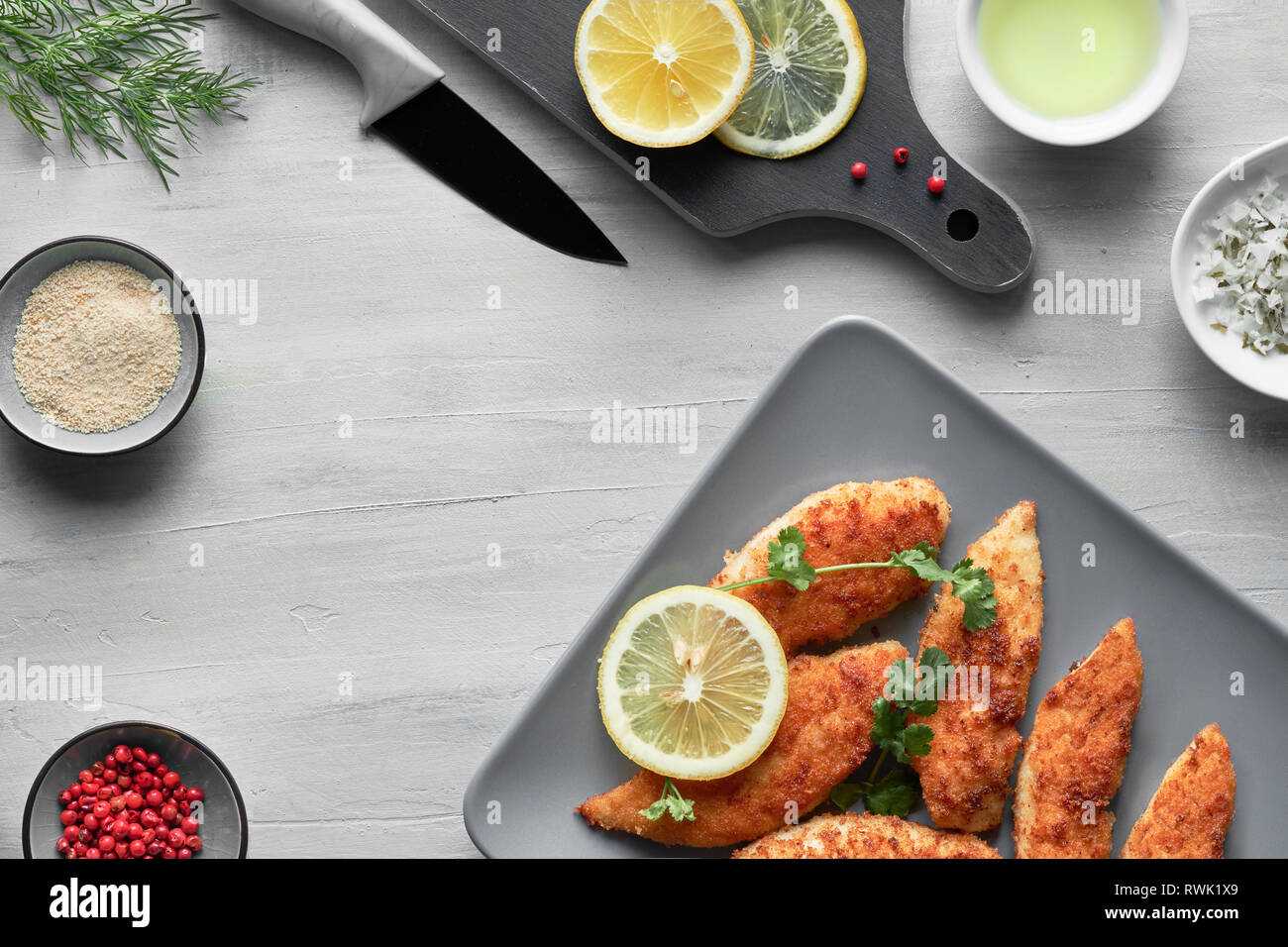 Impanati le strisce di pollo con panatura, olio, sale, pepe rosa, aneto e limone, piatto giaceva su sfondo chiaro Foto Stock