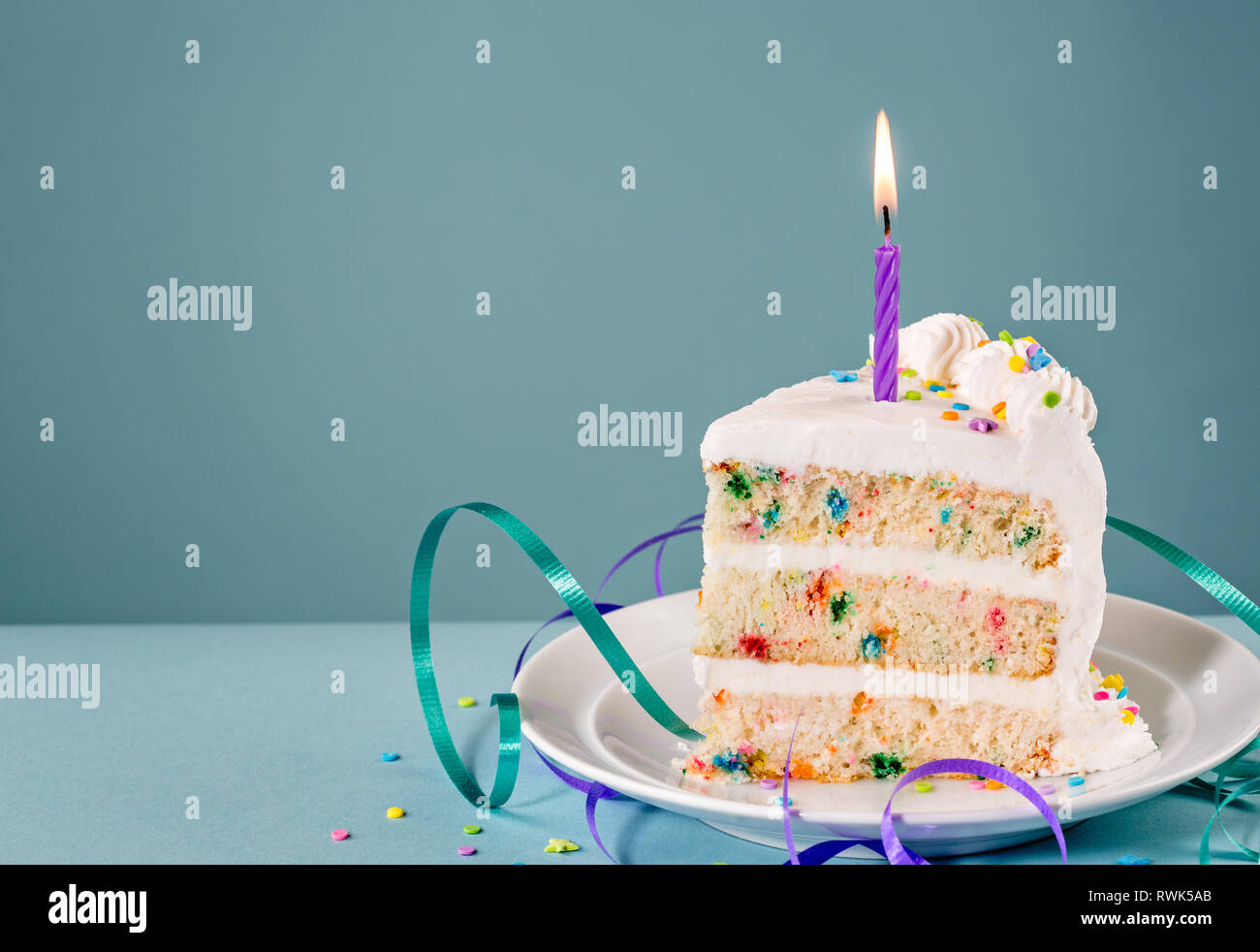 Fetta di torta di compleanno con una candela accesa e nastri su uno sfondo blu. Foto Stock