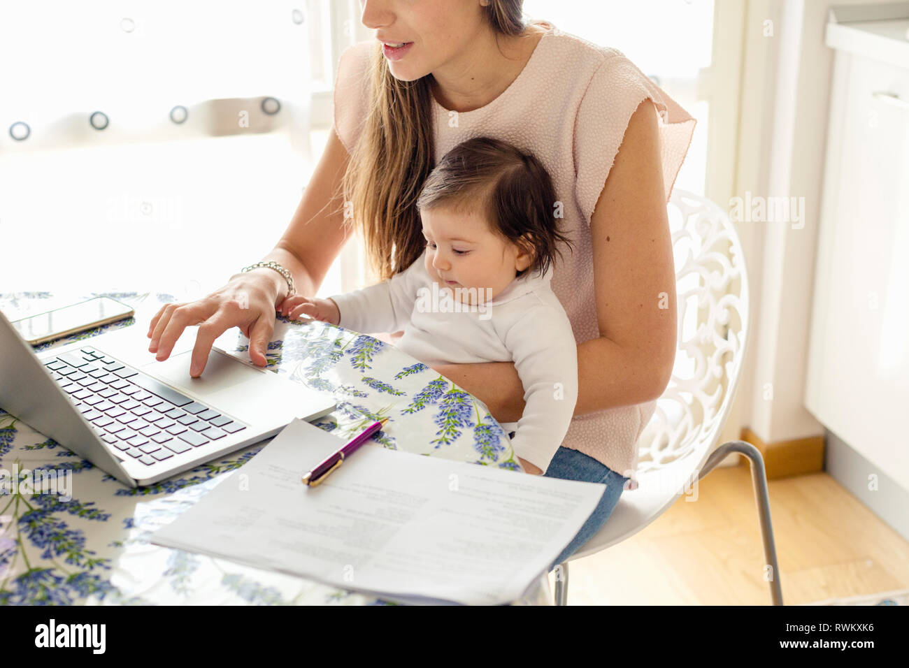 La madre lavora sul computer portatile con la bambina in giro a casa Foto Stock