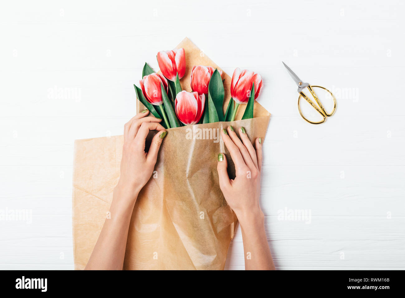 Donna di mani imballaggio bouquet di tulipani rossi in marrone carta eco accanto a forbice bianco sul tavolo di legno, vista dall'alto. Foto Stock