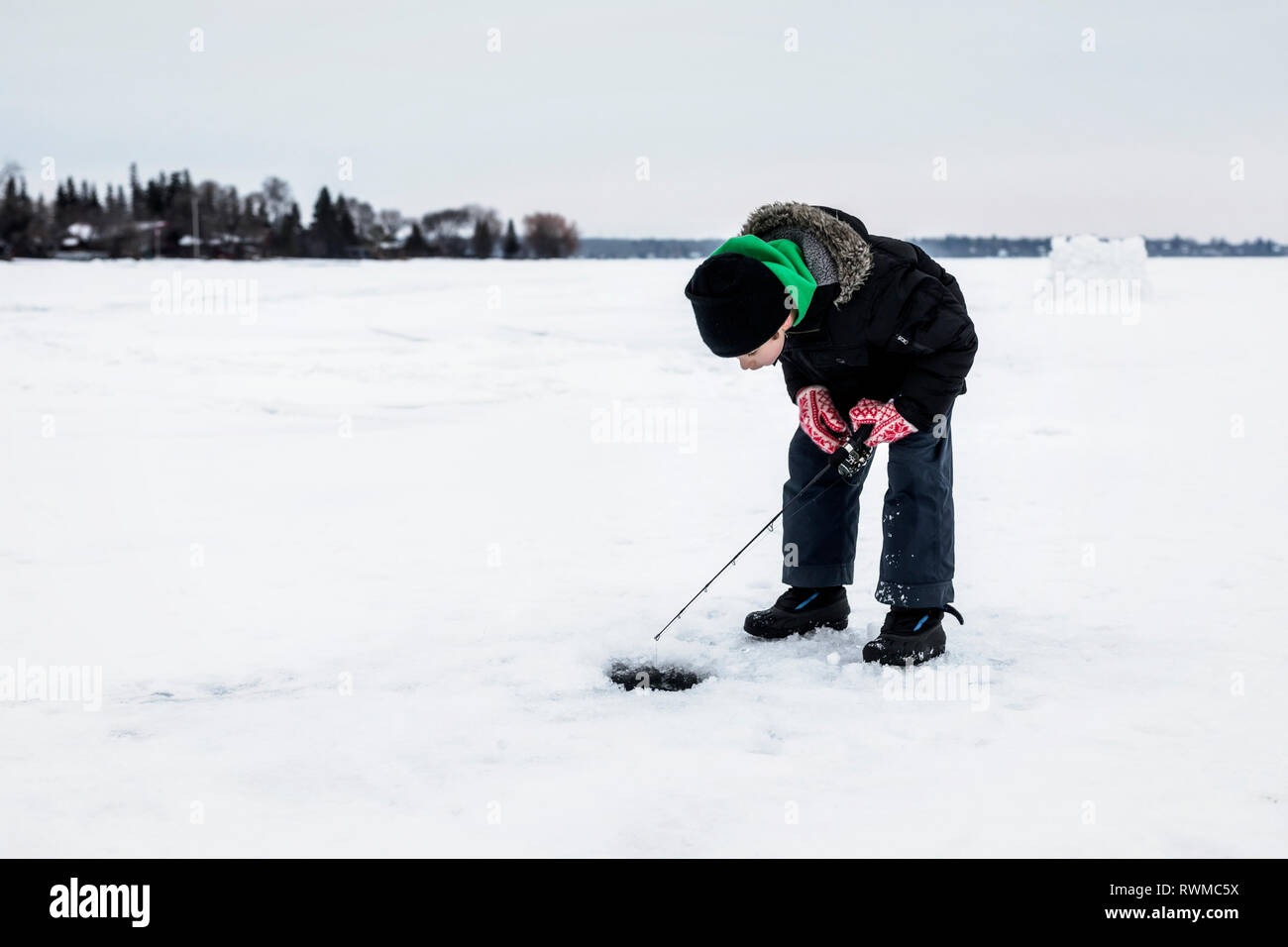 Ragazzo aspettando pazientemente per un morso mentre la pesca sul ghiaccio e guardando verso il basso il foro sul lago Wabamun; Wabamun, Alberta, Canada Foto Stock