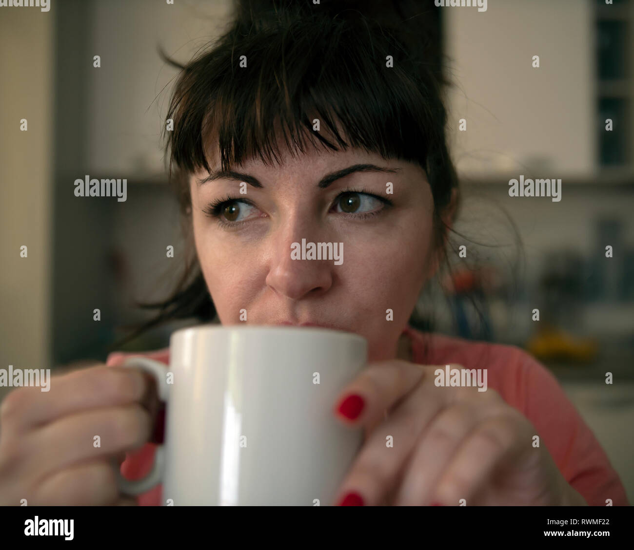 Giovane donna beve caffè al mattino in cucina, stanchi occhi con venature rosse, close-up Foto Stock
