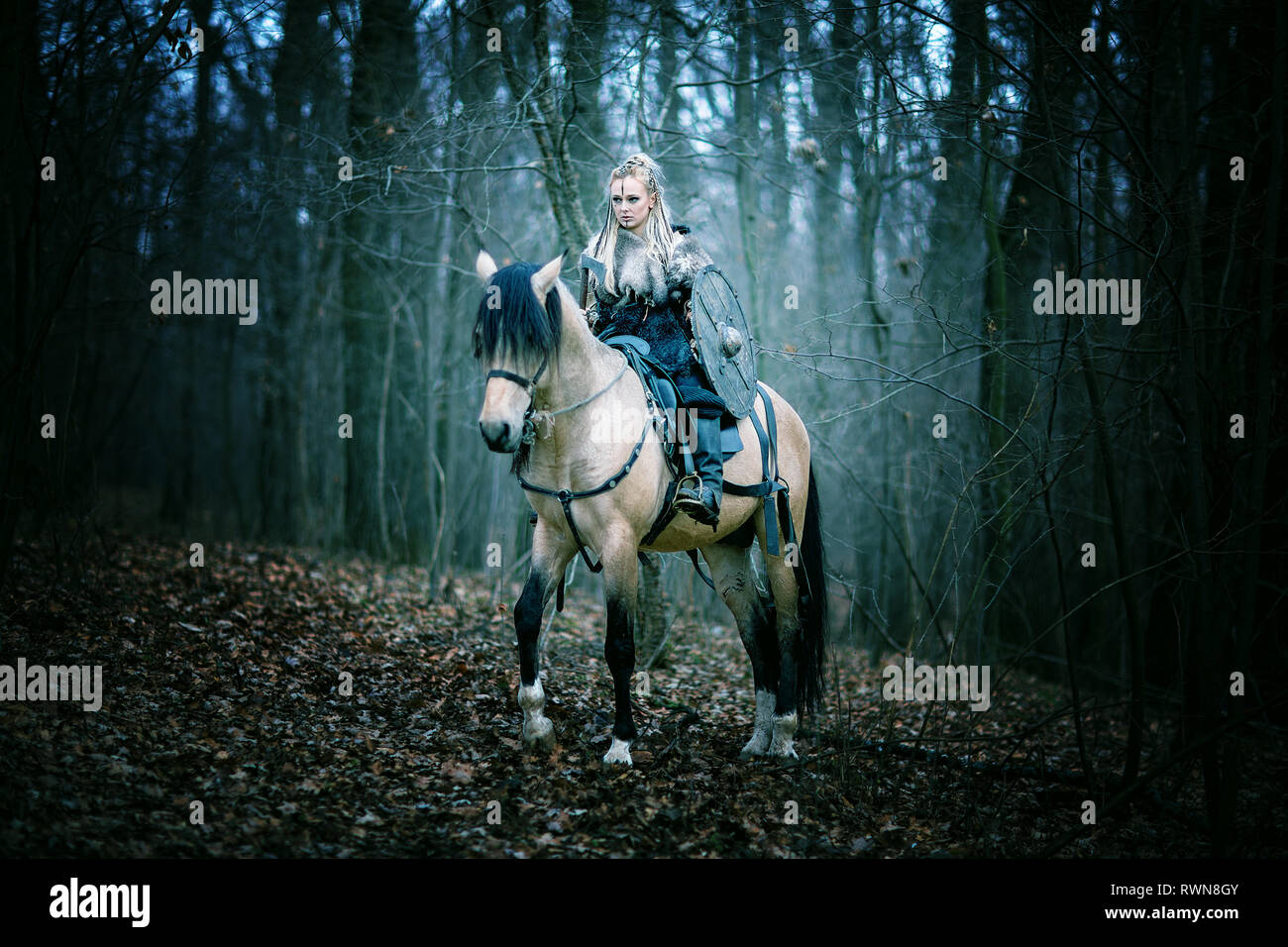 Donna guerriero a cavallo nei boschi. Vichingo scandinavo di equitazione con ax in mano, capelli biondi in abiti tradizionali con collare di pelliccia, guerra mak Foto Stock