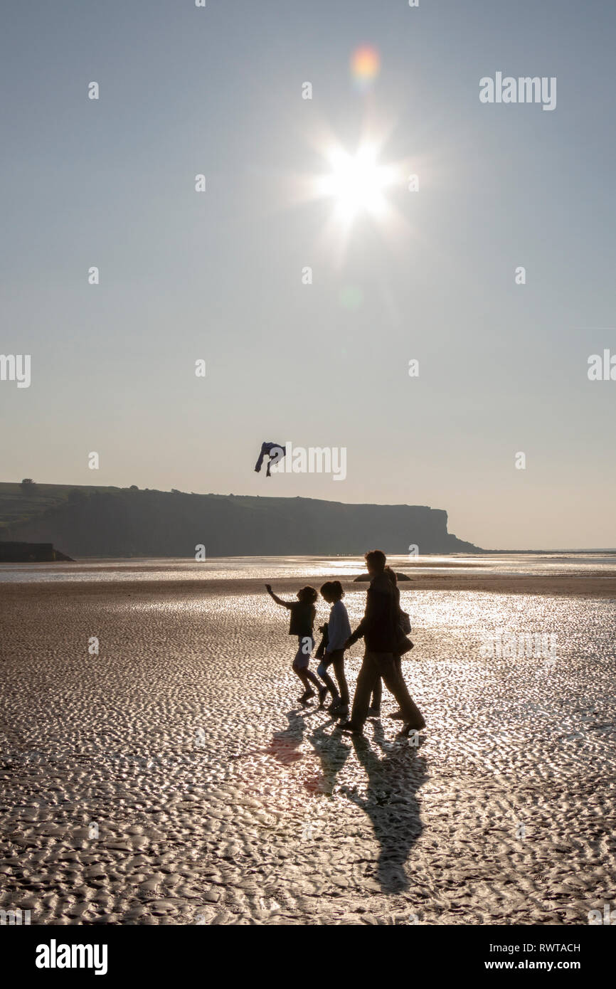Una famiglia attraversando a piedi la spiaggia con la bassa marea su un luminoso sera sull'Arromanches-les-Bains spiaggia al tramonto, Normandia, Francia. Foto Stock