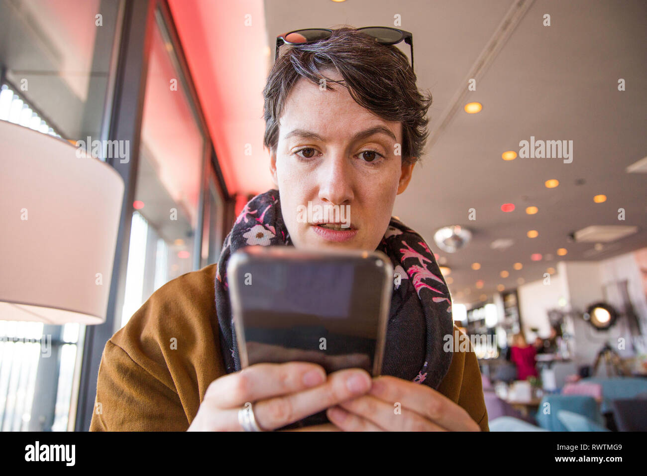 Bella giovane donna in abiti caldi in seduta cafe e controllando il suo smartphone, sorpreso e confuso, sfondo sfocato, testa e spalle Foto Stock