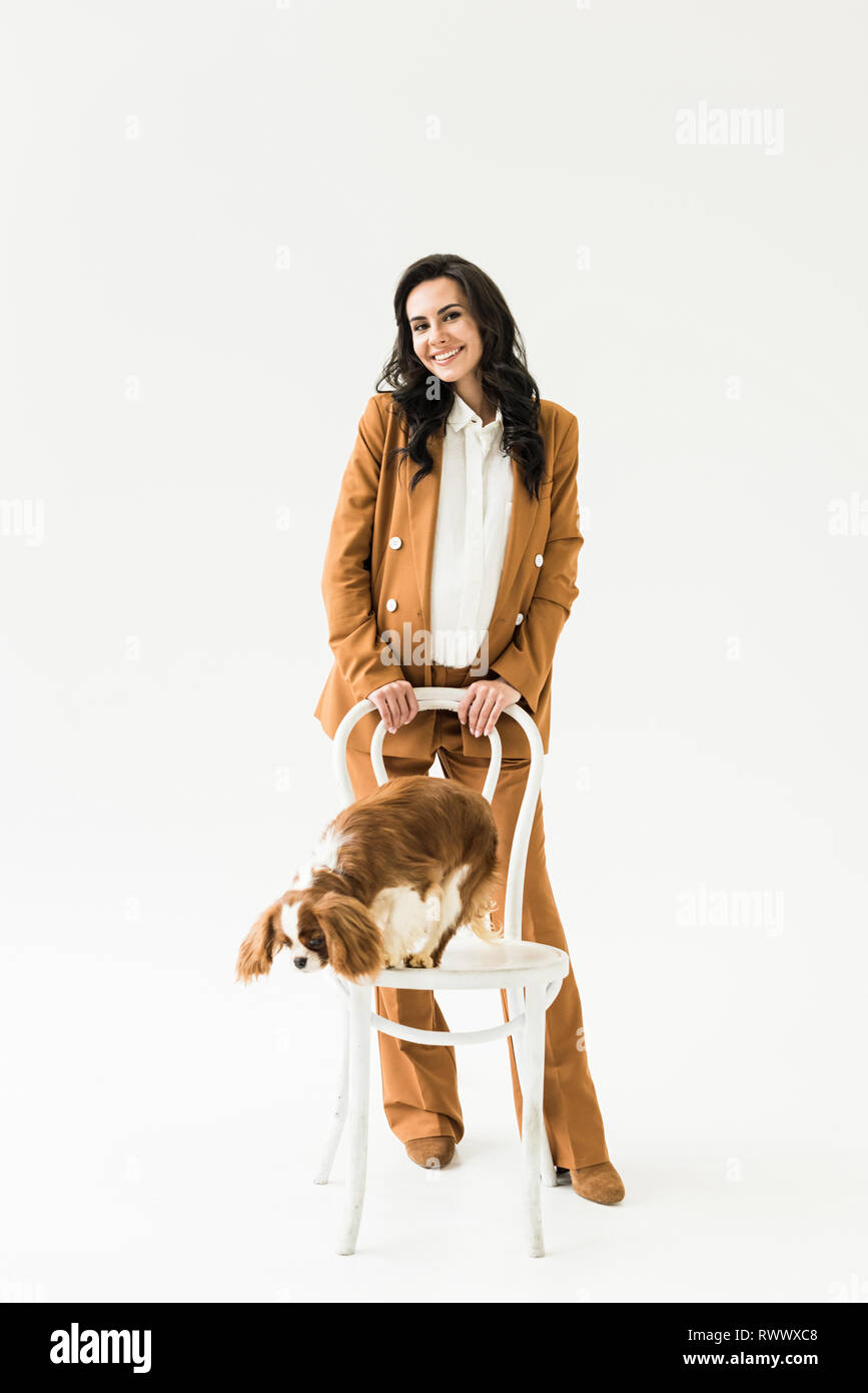 Meravigliosa donna incinta in abito marrone in piedi vicino al cane sulla sedia su sfondo bianco Foto Stock
