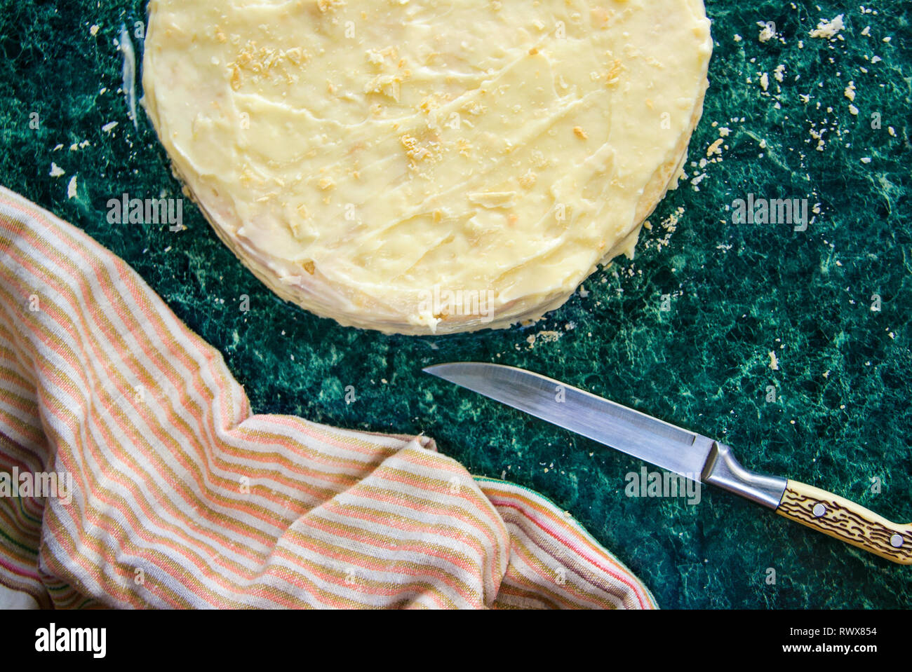 Round layered Napoleone torta con crema pasticcera e cosparso briciole, coltello, asciugamano e mixer su un verde tavola di marmo, vista dall'alto. Il processo di cottura, reci Foto Stock