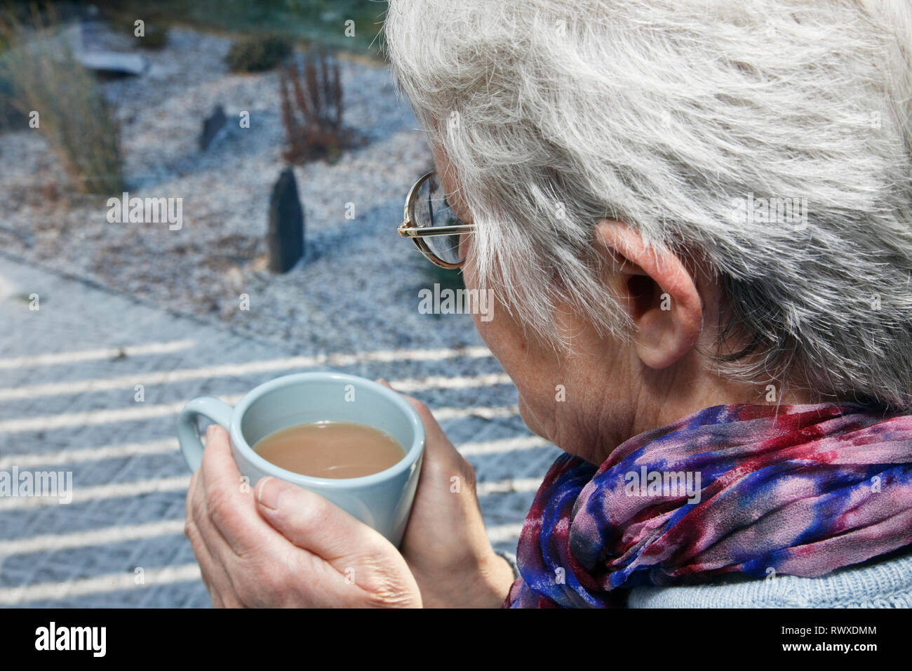 Senior vecchia donna warming mani tenendo una bevanda calda guardando fuori della finestra in un freddo giorno di neve in inverno. Regno Unito, Gran Bretagna Foto Stock