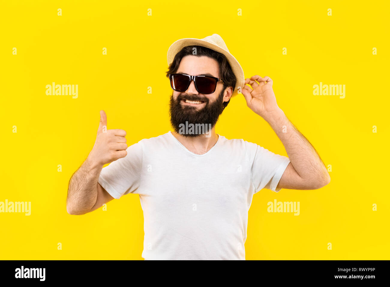 Ritratto in studio di un barbuto ragazzo in una t-shirt e occhiali da sole e panama sorride e mostra un simile concetto di estate spiaggia umore Foto Stock