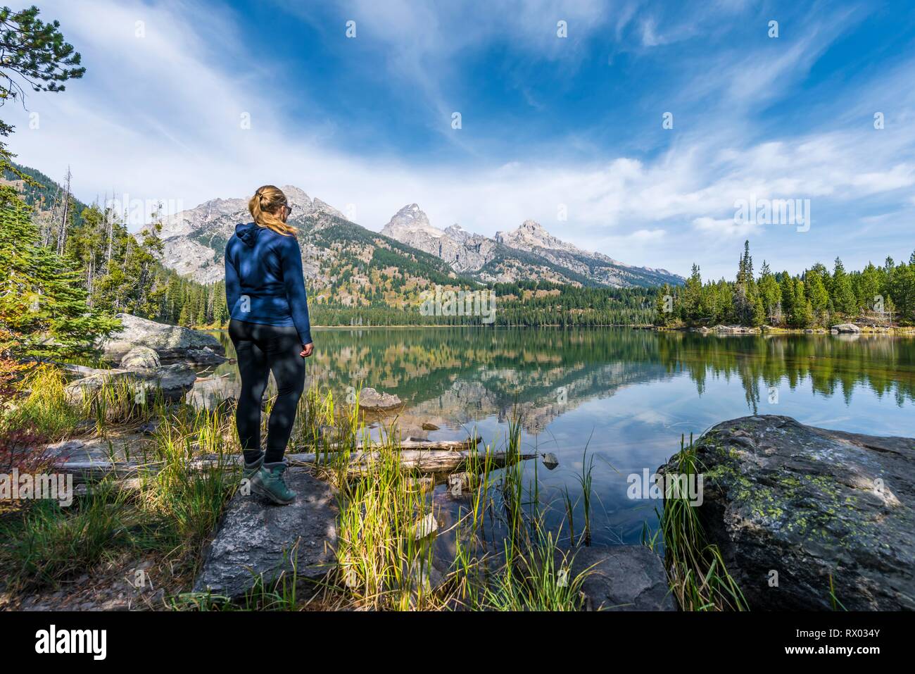 Giovane donna in piedi in un lago e la riflessione in Taggart lago, vista la gamma Teton mountain range, Grand Teton National Park Foto Stock