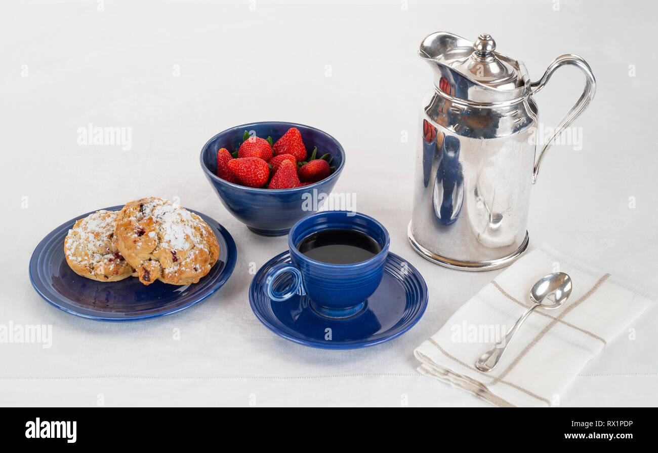 La prima colazione di scones al buio su un piastra di blu e una ciotola di fragole fresche con caffè e un Silver Hotel Servizio in camera caraffa su un lino bianco tovaglia Foto Stock