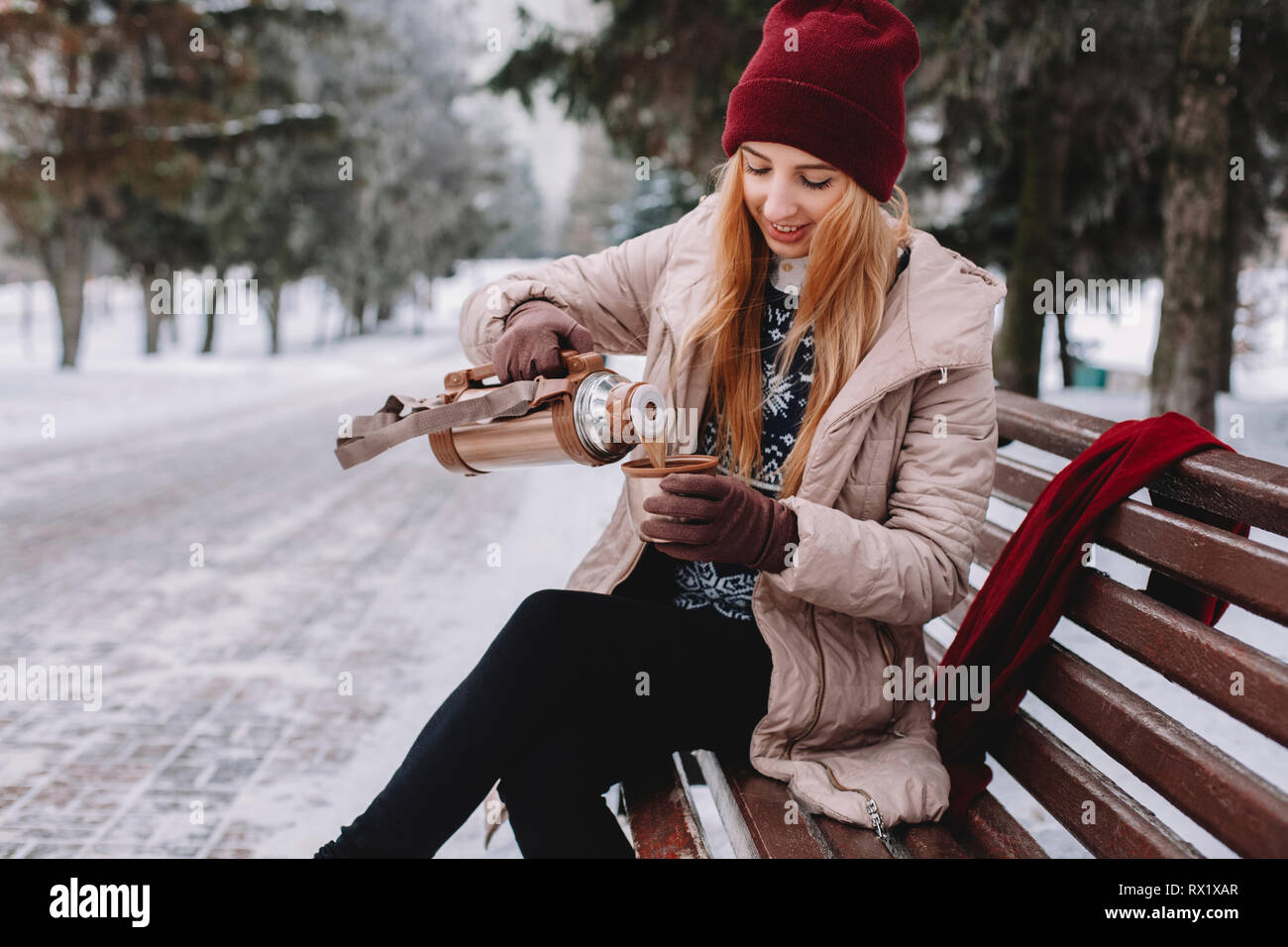 Donna sorridente versando il caffè nella tazza mentre è seduto sulla panchina contro gli alberi al parco durante l inverno Foto Stock