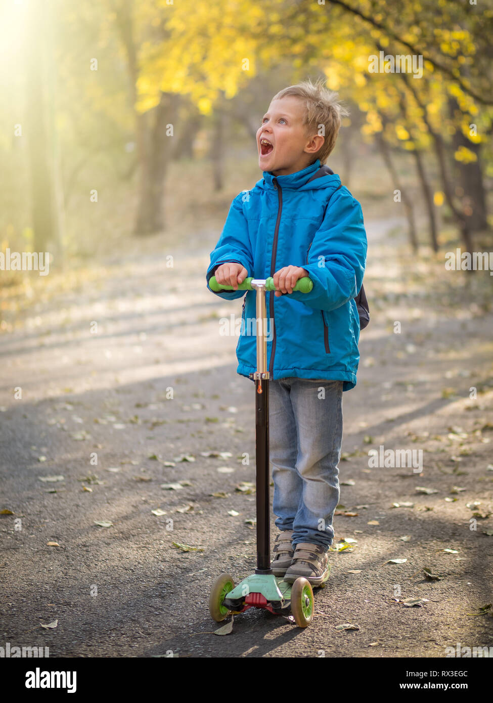 Un ragazzo in blu vestiti equitazione con un ampio sorriso su uno scooter in autunno park. I raggi del sole illuminano la sua faccia. Foto Stock