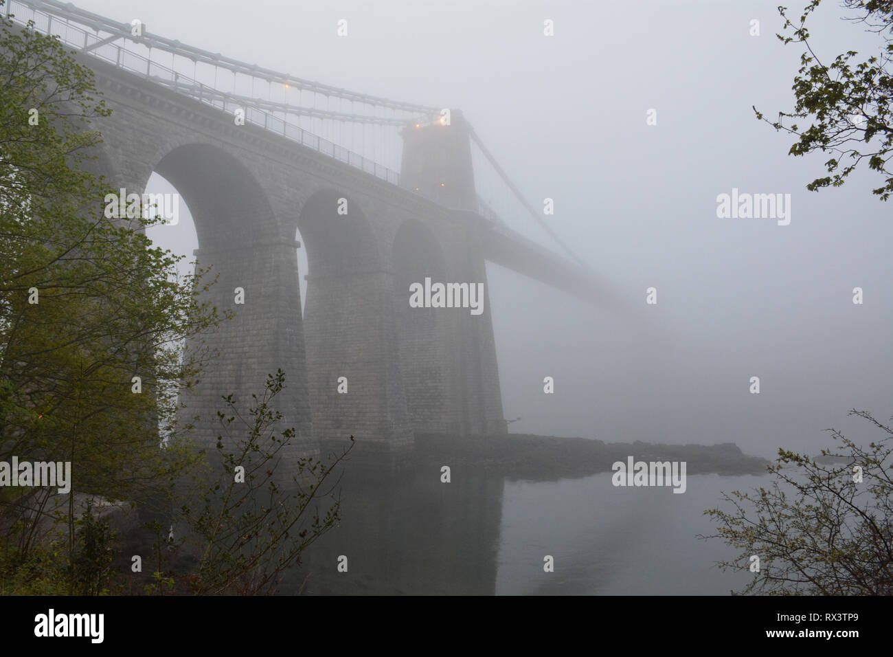 Osservando il Menai Suspension Bridge attraverso la nebbia e la nebbia con il Menai Strait in primo piano Foto Stock