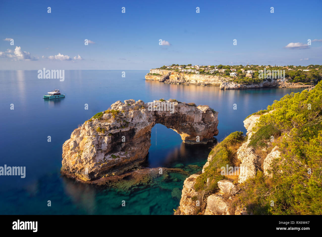 Es Pontas in Mallorca, arco naturale nel Mar Mediterraneo a alba con una barca ancorata nelle vicinanze e ville per vacanze e appartamenti in backgroun Foto Stock