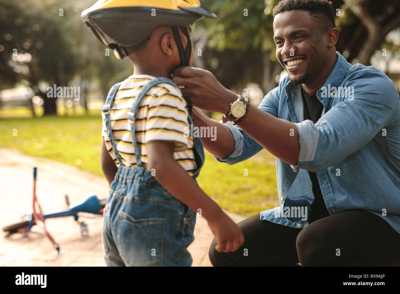 Uomo sorridente aiutando figlio di indossare il casco per andare in bicicletta al parco. Ragazzo pronte da indossare il casco protettivo per avviare il ciclismo. Foto Stock