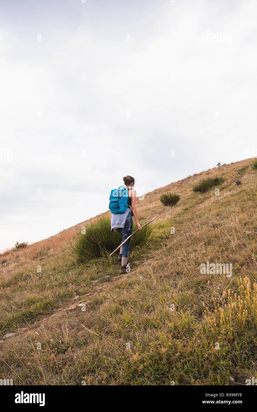 Vista posteriore di un escursionista femmina con zaino camminando sulla montagna erbosa contro sky Foto Stock