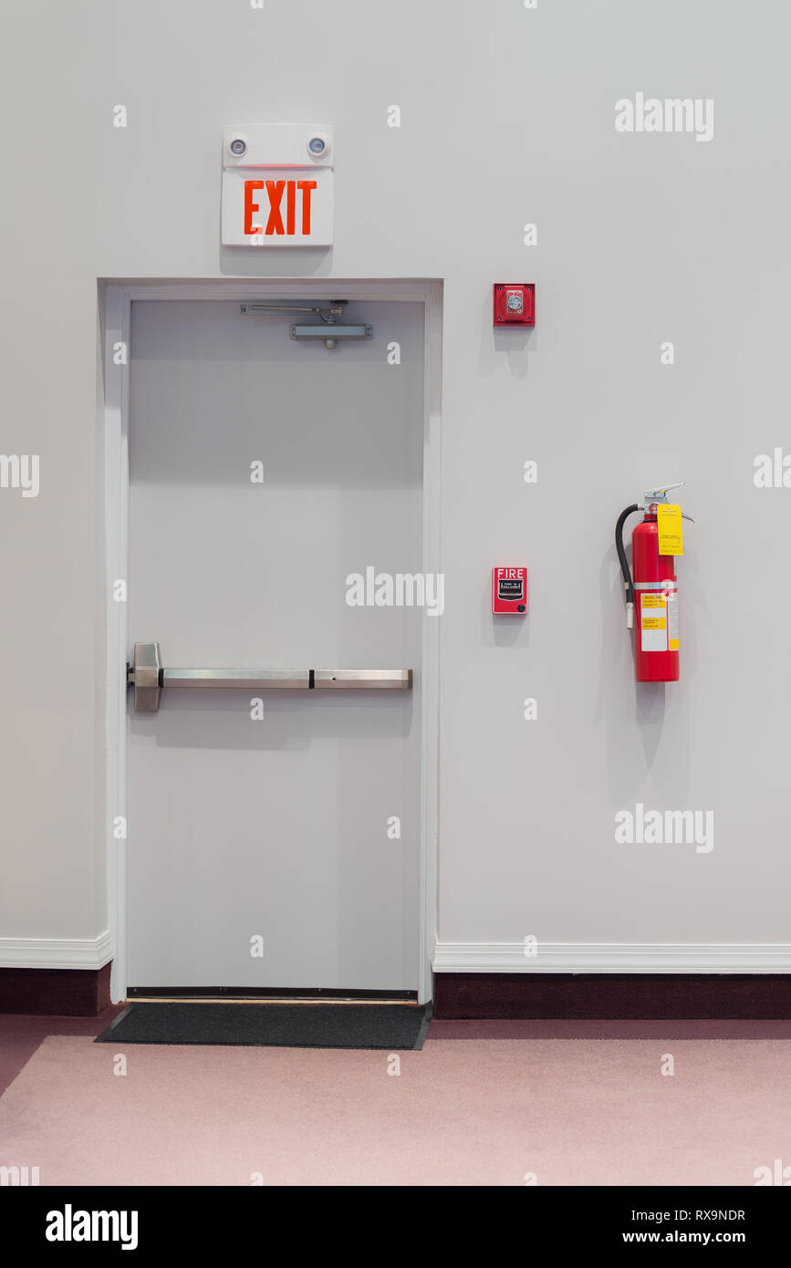 Colpo verticale di un uscita di emergenza porta con un allarme incendio, estintore, e di una luce di emergenza appeso alla parete accanto ad essa. Foto Stock