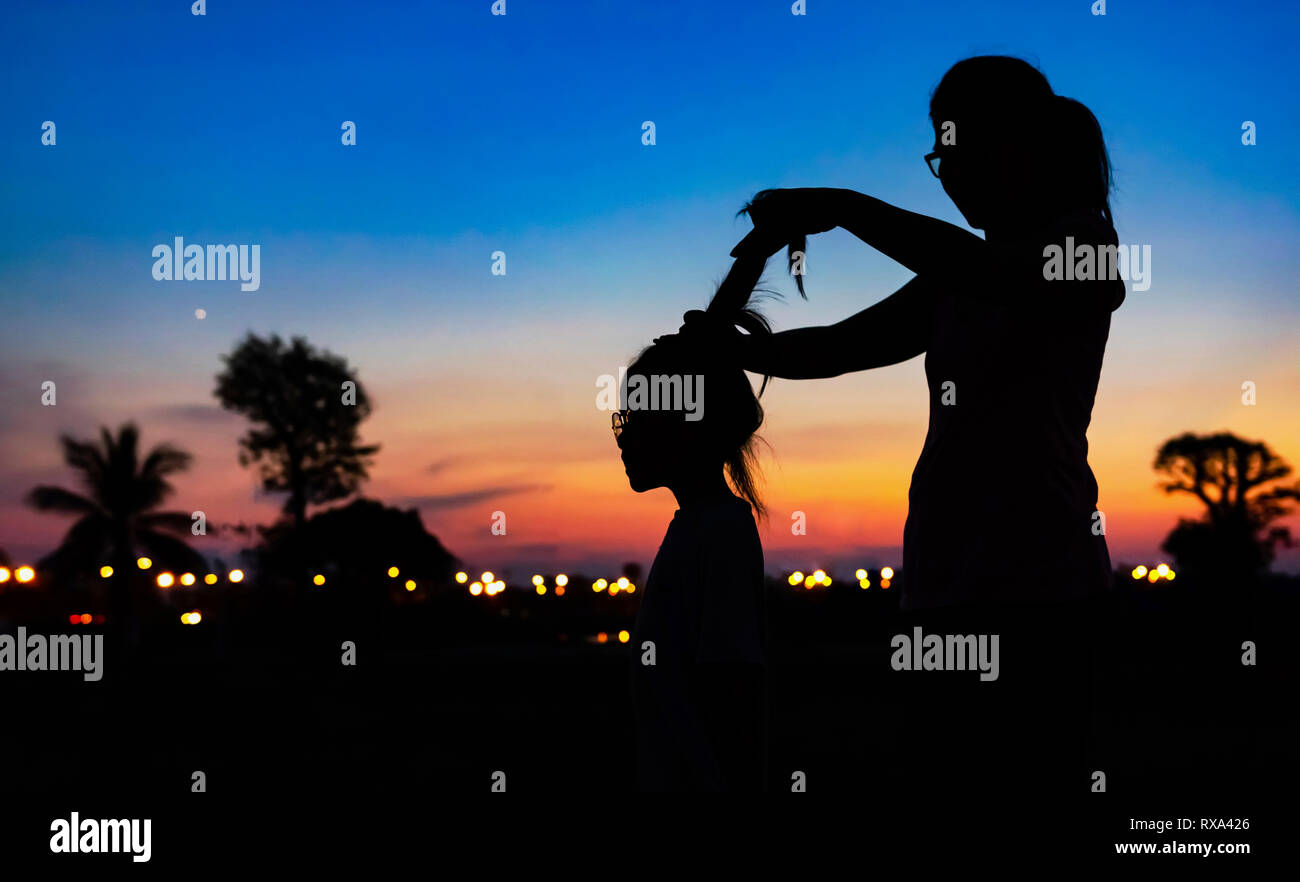 Vista laterale di silhouette madre figlia di legatura di capelli contro il cielo drammatico durante il tramonto Foto Stock