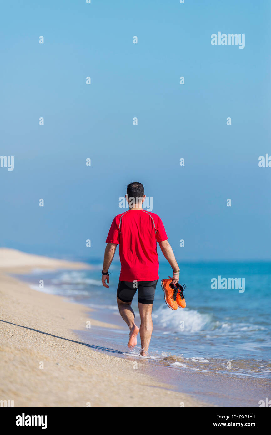 Vista posteriore dell'uomo azienda sport scarpe camminando sulla spiaggia a spiaggia contro sky Foto Stock