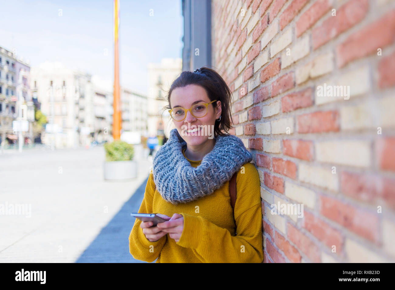 Ritratto di sorridente turista femminile di indossare un abbigliamento caldo utilizzando smart phone mentre in piedi contro la parete in città Foto Stock