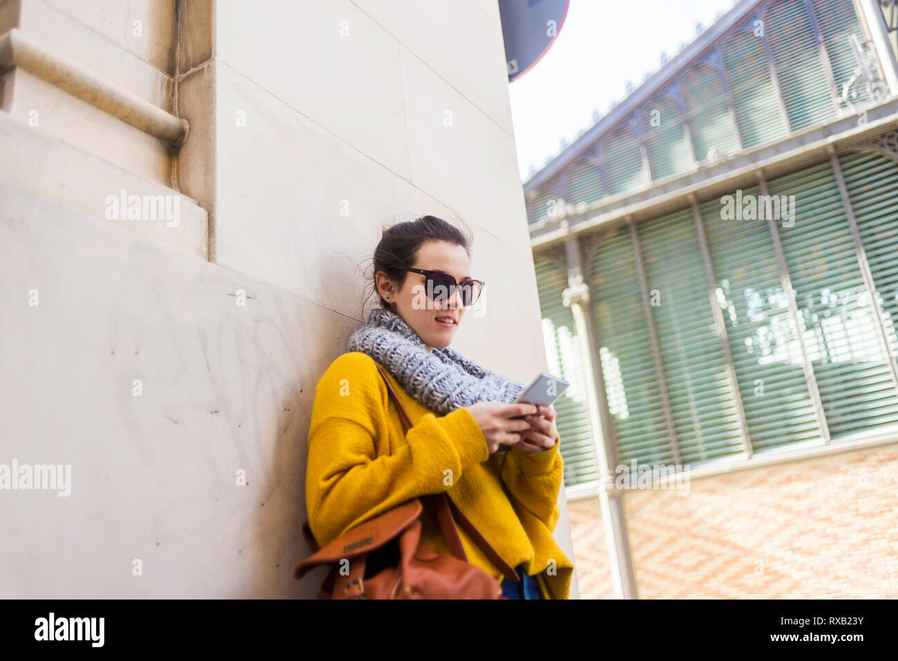 Turista femminile di indossare un abbigliamento caldo e occhiali da sole utilizzando smart phone mentre in piedi contro la parete Foto Stock