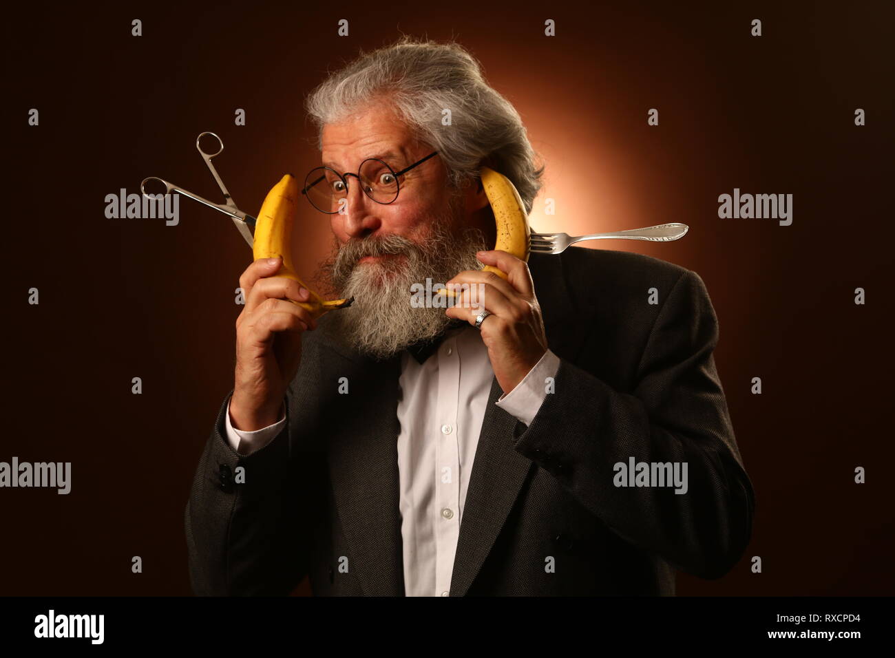 Ritratto di un grey-haired anziano gentiluomo con un shaggy i baffi e barba, in una camicia bianca e giacca grigia, indossare occhiali e una forcella, tenendo un Foto Stock