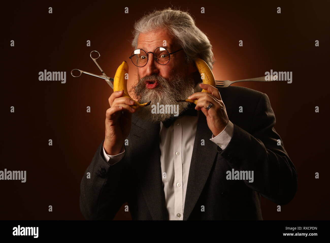 Ritratto di un grey-haired anziano gentiluomo con un shaggy i baffi e barba, in una camicia bianca e giacca grigia, indossare occhiali e una forcella, tenendo un Foto Stock