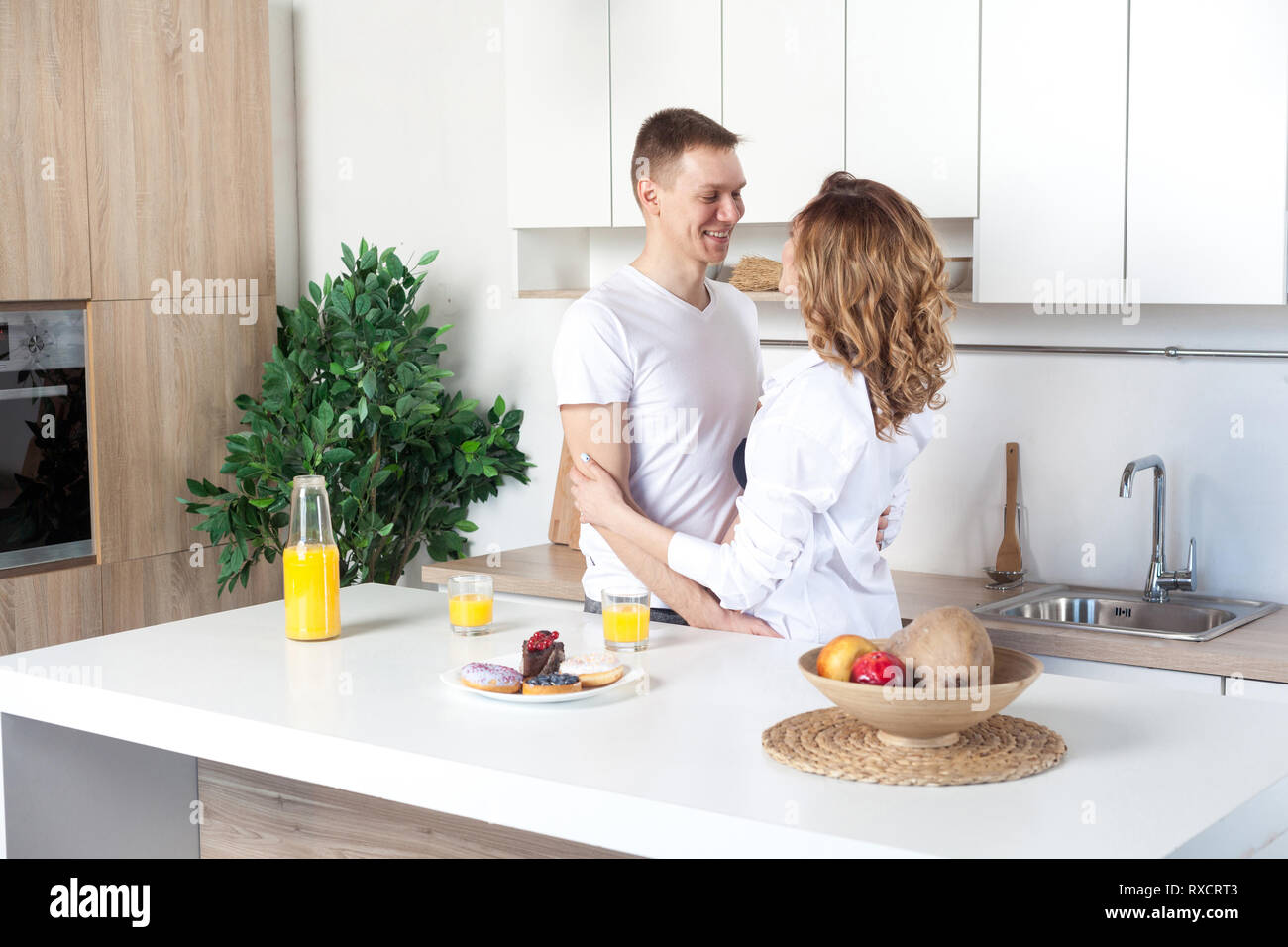 Giovani felice coppia sposata abbraccia in piedi vicino a tavola in cucina. Il marito abbraccia la sua moglie incinta, ai genitori di godere di ogni altro guardando agli occhi. Amore Foto Stock