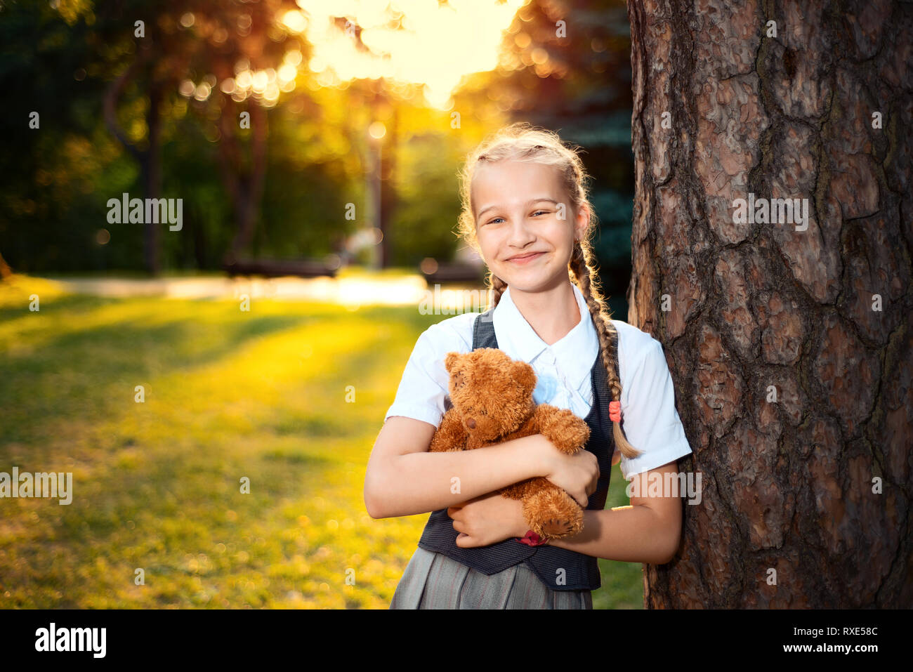 Ritratto di schoolgirl con pig-tail in uniforme al tramonto. lo studente si erge nei pressi di un albero e detiene un soft bear toy Foto Stock
