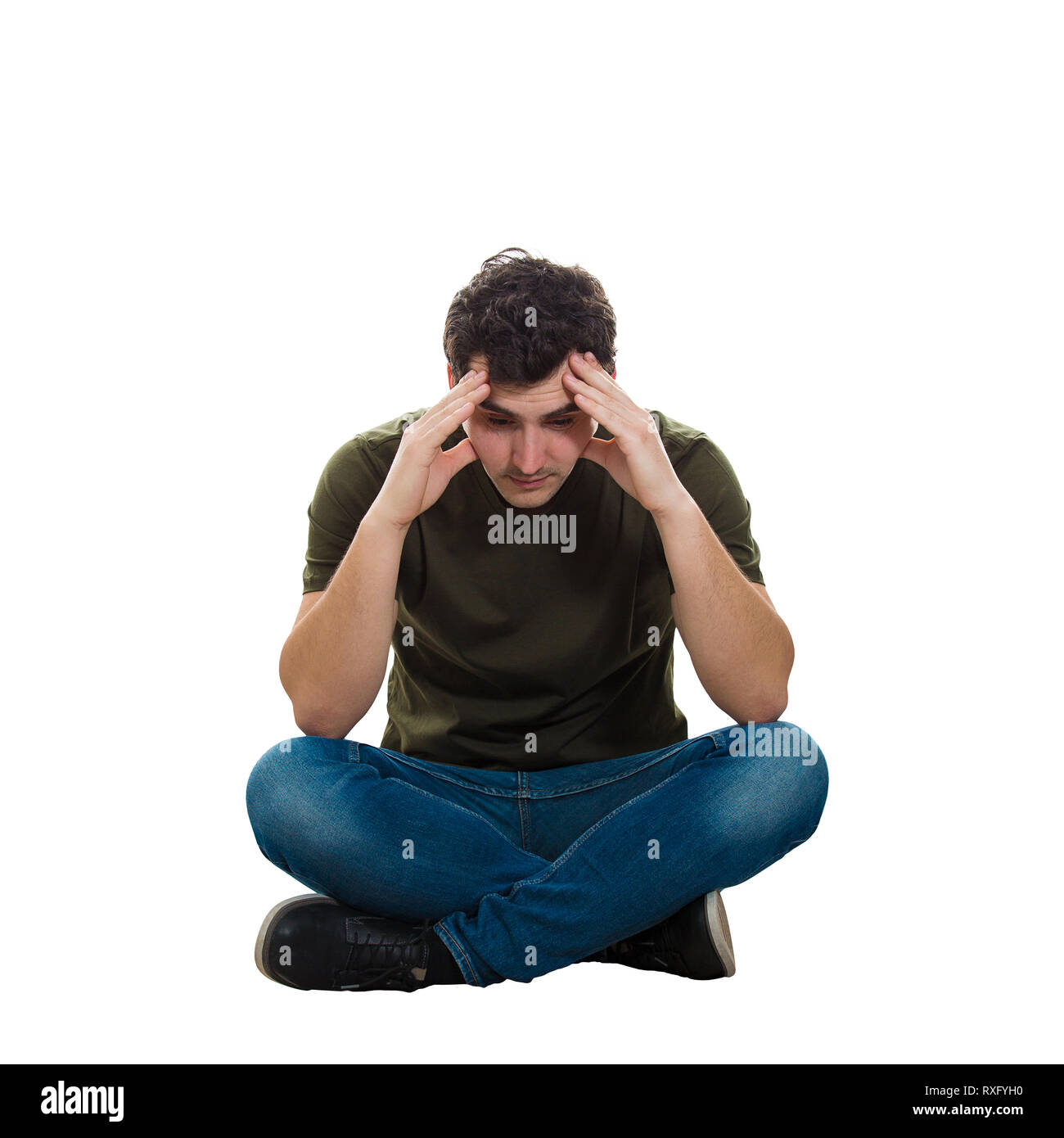Arrabbiato e stanco giovane uomo casual da indossare jeans e maglietta, seduto sul pavimento con le gambe incrociate, puntando le mani per la fronte guardando giù riflessivo Foto Stock