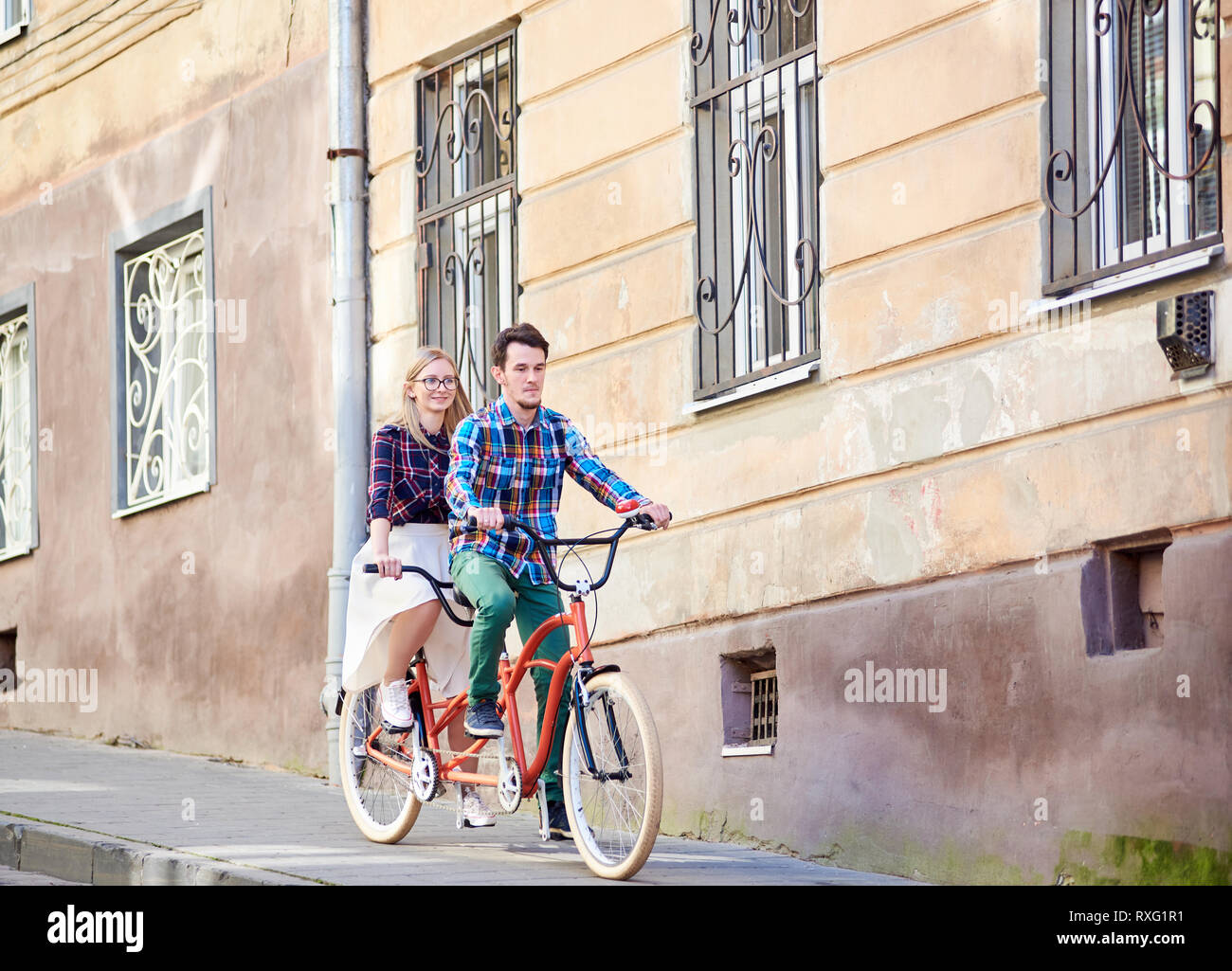 Bel giovane uomo barbuto e bella donna bionda in bicchieri in sella insieme a doppio tandem bicicletta lungo il marciapiede pavimentato su soleggiate giornate estive da Foto Stock