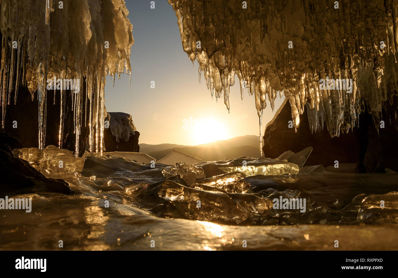 Incredibile sunrise oltre le montagne e il lago ghiacciato in inverno dalla caverna di ghiaccio con ghiaccioli. Lago Teletskoye, Altai, Russia Foto Stock