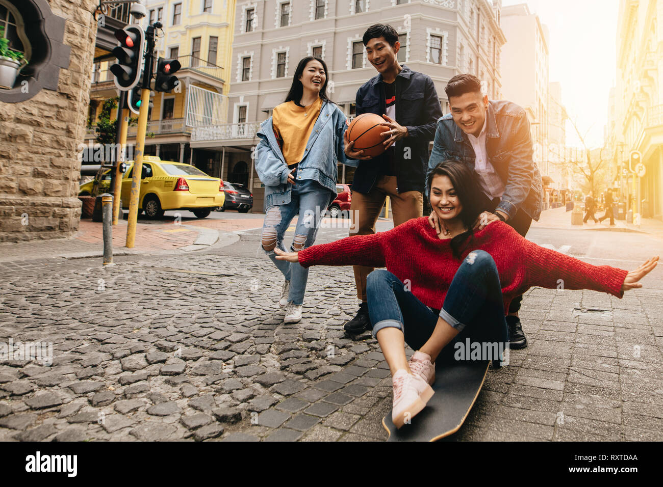 Asian giovani nella città con lo skateboard e il basket. Donna essendo spinto da uomo su skateboard all'aperto sulla strada con i loro amici a piedi Foto Stock