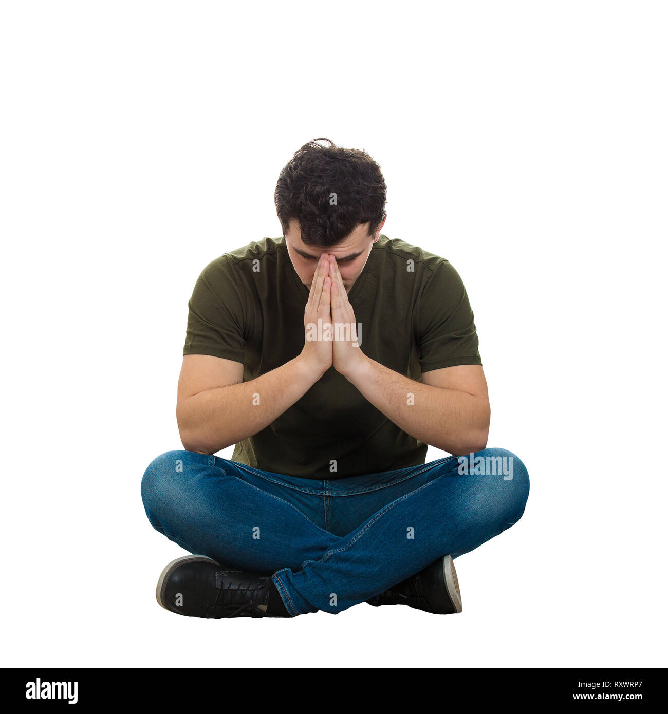 Sottolineato casual giovane uomo seduto sul pavimento con le mani piegate insieme e gambe incrociate guardando in giù come pregare isolate su sfondo bianco. P Foto Stock
