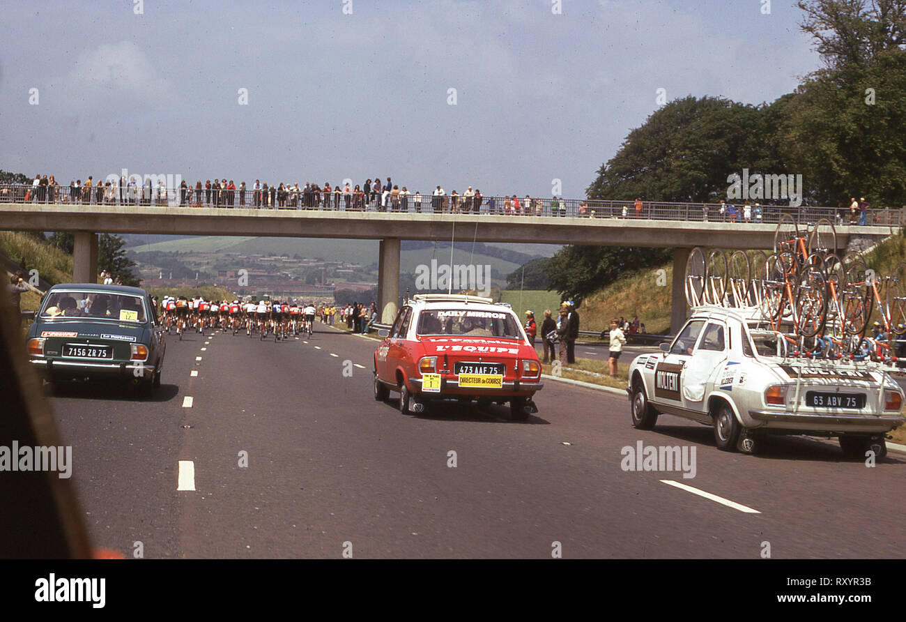 1974 Tour de France, tappa britannica, ciclisti risalendo il Plymouth by-pass, seguita dalla pressa e supporto di vetture Peugeot e guardati da spettatori su un ponte. Foto Stock