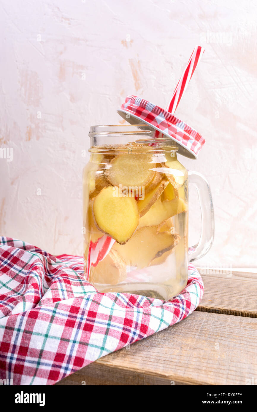 Vaso di vetro con acqua allo zenzero con cannuccia e tartan rosso cappuccio rosso sul panno a scacchi su tavole di legno su sfondo chiaro. Foto Stock