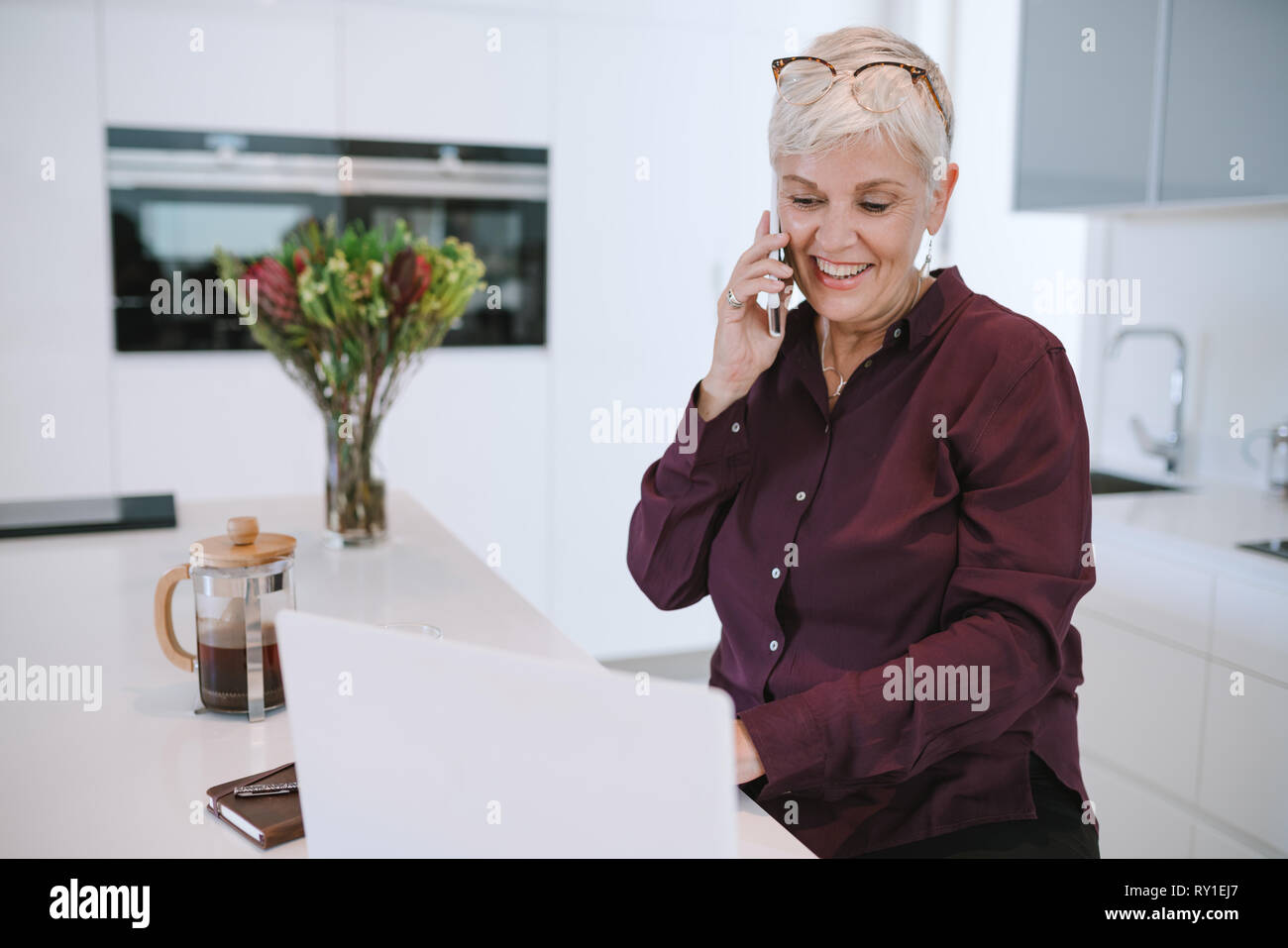 Sorridente casual donna senior utilizzando laptop mentre si parla sullo smartphone. Felice donna matura lavora con un telefono cellulare e il computer portatile a casa. Business donna u Foto Stock