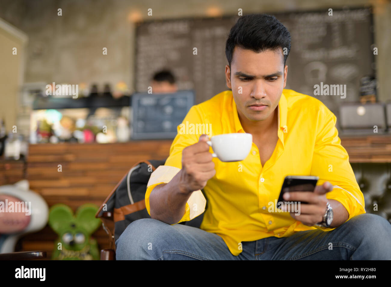 Bel giovane uomo indiano tramite telefono e seduta al coffee shop Foto Stock