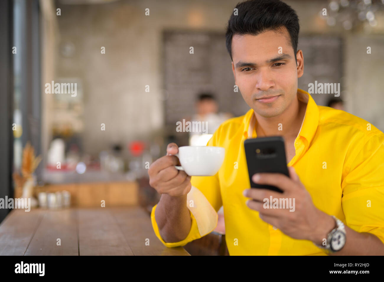 Bel giovane uomo indiano tramite telefono e seduta dalla finestra al cafe Foto Stock